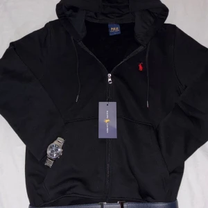 Svart Ralph Lauren full zip hoodie  - Svart hoodie från Ralph Lauren med dragkedja och huva. Klassisk röd logga broderad på bröstet, snörning i huvan och känguruficka framtill. Tillverkad i mjuk bomull, perfekt för chill dagar. Snygg och enkel att matcha med jeans eller joggers.