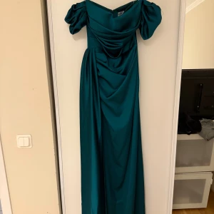 Grön långklänning i satin  - Elegant smaragdgrön långklänning köpt utomlands i glansig satin. Klänningen har v-ringad draperad överdel, markerad midja och puffiga korta ärmar. Långt fall med snygg slits i sidan. Perfekt för dig som vill sticka ut med en lyxig look.