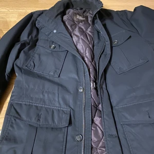 Svart parkajacka Oscar Jacobson stl 50 - Parkajacka från Oscar Jacobson i blå med quiltat foder och flera praktiska fickor framtill. Jackan har dragkedja och knappar, samt hög krage. Perfekt för kyligare dagar och har en clean, stilren look.