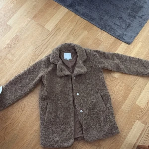 Brun teddyjacka från H&M - Mysig brun teddyjacka från H&M med klassisk krage och knäppning framtill. Jackan har två fickor på sidorna och är fodrad på insidan för extra komfort. Perfekt för kyliga dagar och ger en cool, avslappnad vibe.