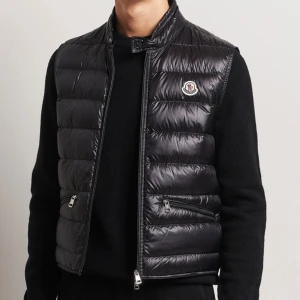 Svart dunväst från Moncler - Snygg svart dunväst från Moncler med glansig finish och quiltad design. Västen har hög krage, dragkedja framtill och två fickor med dragkedja. Ikonisk Moncler-logga på bröstet ger en exklusiv touch. FINNS I FLERA STORLEKAR 