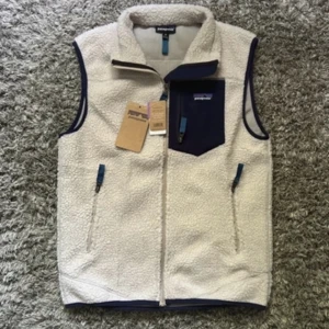 Vit fleeceväst från Patagonia - Säljer en vit fleeceväst från Patagonia med marinblå detaljer. Västen har hög krage, dragkedja framtill och en snygg bröstficka med dragkedja. Perfekt för lager-på-lager och outdoor-vibes. Klassisk Patagonia-logga på bröstet.