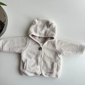 Vit teddyjacka med öron  - Supersöt vit teddyjacka från H&M i storlek 56. Jackan har huva med små öron, dragkedja framtill och två fickor. Insidan av huvan är randig i beige och vitt. Perfekt för att hålla de minsta varma och mysiga.