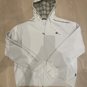 Vit Burberry hoodie med rutig huva XL - Exklusiv vit hoodie från Burberry i storlek XL med klassiskt rutigt foder i huvan. Tröjan har dragkedja framtill, snörning vid huvan och subtil logga på bröstet. Perfekt för dig som vill ha en stilren och lyxig streetwear-look.