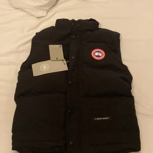 Svart dunväst från Canada Goose - Svart dunväst från Canada Goose med klassisk logga på bröstet och två stora fickor framtill. Västen har hög krage, tryckknappar och är fodrad för extra värme. Perfekt för lager-på-lager och har en clean, stilren look.
