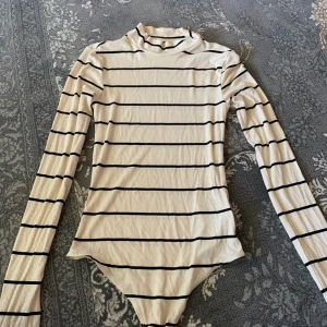 Randig långärmad polotopp - Snygg långärmad polotopp i vitt med svarta horisontella ränder. Toppen har en hög krage och är figurnära med ribbad struktur. Perfekt att styla med jeans eller kjol för en trendig look.