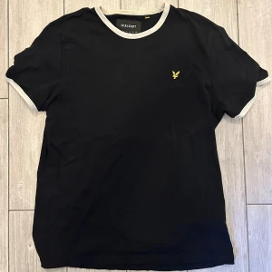 Svart t-shirt från Lyle & Scott - Svart t-shirt från Lyle & Scott med gråa ribbade kanter vid hals och ärmslut. Klassisk passform och liten gul logga broderad på bröstet. Perfekt basic-plagg med stilren look. Skriv för fler bilder!
