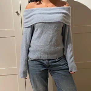 Ljusblå offshoulder stickad tröja - Superfin ljusblå stickad tröja med offshoulder-design och långa ärmar. Tröjan har bred krage som vilar snyggt över axlarna och ribbade muddar vid ärmslut och nederkant. Perfekt för dig som vill ha en trendig och mysig look.