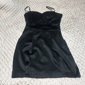 Svart kort klänning från Lindex - Supersnygg svart kort klänning från Lindex med smala axelband och markerad midja. Klänningen har en lätt glansig yta och dragkedja i sidan. Perfekt för dig som gillar en stilren och elegant look.