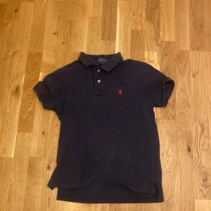 Mörkblå pikétröja från Ralph Lauren - Säljer en mörkblå pikétröja från Polo Ralph Lauren i slim fit-modell. Tröjan har klassisk krage, korta ärmar och knäppning med två knappar. Ikoniska röda loggan broderad på bröstet. Perfekt för en clean och stilren look. Vid snabb affär kan priset ändras