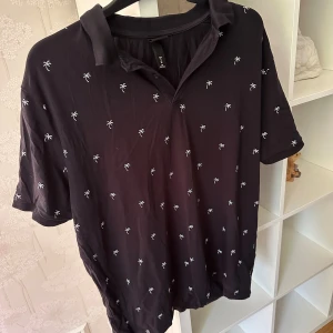 Svart kortärmad pikétröja med palmmönster strl: XL - Svart pikétröja från H&M med korta ärmar och krage. Tröjan har små vita broderade palmer över hela tyget och är tillverkad i mjuk bomull. Perfekt för en chill sommarlook.