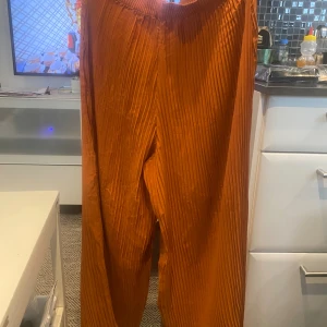Plisserade orange vida byxor - Snygga plisserade byxor i en stark orange färg med bred resår i midjan. Byxorna har en loose och wide passform med raka ben och är tillverkade i ett lätt, glansigt material som ger ett flowy intryck. Perfekta för dig som vill sticka ut med färg och stil.