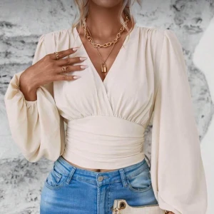 Beige omlottopp med ballongärmar - Säljer en beige omlottopp med djup v-ringning och bred, markerad midja. Toppen har långa ballongärmar med resår vid handlederna och smock baktill i midjan för en snygg passform. Perfekt att styla med jeans för en trendig look.