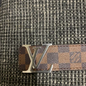 Louis Vuitton rutigt brunt bälte - Snyggt bälte från Louis Vuitton med klassiskt LV-spänne i silverfärgad metall. Bältet har ett rutigt mönster i mörkbrunt och ljusbrunt och är gjort i skinn. Perfekt accessoar för att lyfta din outfit med en lyxig touch.