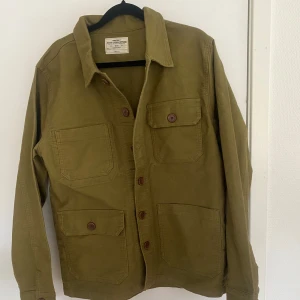 Olivgrön overshirt från Master Clothing - Olivgrön overshirt från Master Clothing Suppliers i 100% bomull. Jackan har fyra stora fickor framtill, knäppning med bruna knappar och klassisk krage. Perfekt lager-på-lager-plagg med avslappnad vibe.