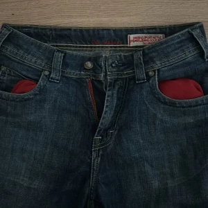 Blå lågmidjade jeans med broderade bakfickor - Säljer ett par blå jeans från 2000s talet med raka ben och snygga broderade detaljer på bakfickorna. Jeansen har röda innerfickor fram och kontrastsömmar. Väldigt små och tajta på mig, och lite kort på mig som gillar när dem går över foten. Jag är 158 cm. Rekommenderar byxorna för dem som är lite kortare 