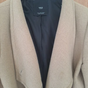 Beige kappa med bälte från MNG - Snygg beige kappa från MNG med bälte i midjan och stora slag. Lång modell med fickor på sidorna och helsvart foder. Perfekt för dig som vill ha en stilren och klassisk look under höst och vinter.