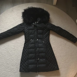 Svart lång Rockandblue dunjacka - Snygg svart lång dunjacka från Rockandblue med quiltade partier och stor huva med fuskpäls. Jackan har dragkedja, knappar och flera fickor med dragkedja. Perfekt för kalla dagar och har en figurnära passform. Passar storlek S/36 också 💕