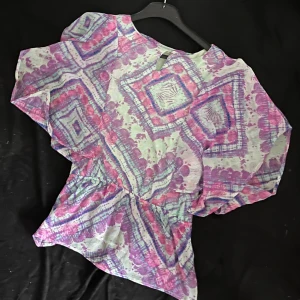 Ljus blus med lila och rosa mönster - Säljer en luftig blus med vida ärmar och resår i midjan. Blusen har ett färgstarkt geometriskt mönster i lila, rosa och blått på ljus botten. Perfekt för dig som gillar boho-vibes och vill sticka ut med färg och form.