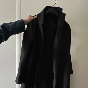 Svart lång kappa i ull - Stilren svart kappa med rak passform och klassisk krage. Jackan är lång och tillverkad i ett mjukt ullmaterial, perfekt för kyligare dagar. Enkel och tidlös design som passar till många olika outfits.