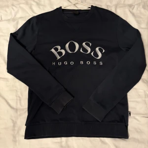 Hugo Boss sweatshirt  - Snygg mörkblå sweatshirt från Hugo Boss med stort broderat BOSS-tryck på bröstet. Tröjan har rund hals, långa ärmar och ribbade muddar vid ärmslut och nederkant. Har små defekter, syns på bilderna där jag använd ficklampa, inte synliga annars. 