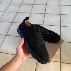 Svarta sneakers från Christian Louboutin - Snygga svarta sneakers i mocka från Christian Louboutin med klassisk röd sula och signatur på undersidan. Skorna har rund tå, platt sula och svarta snören. Insidan är beige och de har en stilren, minimalistisk design som passar till allt.