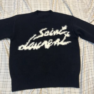 Svart Saint Laurent stickad tröja - Svart stickad tröja från Saint Laurent med vit, fluffig logga framtill i cool graffiti-stil. Klassisk rund halsringning och ribbade muddar vid ärmslut och nederkant. Perfekt för dig som vill ha en statement piece med lyxig känsla.