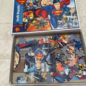 Trefl Puzzle Superman 7+ - Superman-pussel från Trefl, rekommenderas för barn från 7 år. Färgglada bitar med motiv från DC Comics. Pusslet är delvis lagt och bitarna ser hela ut. Perfekt för fans av superhjältar och pussel.