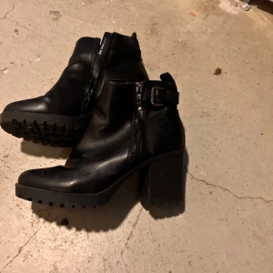 Svarta boots med klack och spänne - Snygga svarta boots i skinn med grov sula och rejäl klack. Dragkedja på sidan och dekorativt spänne vid ankeln ger en cool look. Perfekta för dig som vill ha både stil och edge. Passar till jeans eller kjol för en trendig outfit.