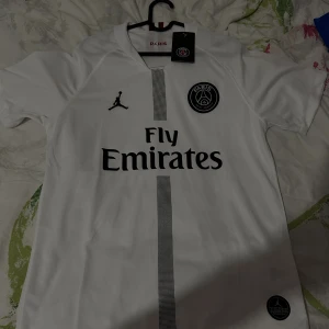 Vit PSG fotbollströja Jordan M - Snygg vit PSG fotbollströja i samarbete med Jordan. Tröjan har svart Paris Saint-Germain-märke på bröstet, Fly Emirates-tryck och diskret rand i mitten. Tillverkad i lätt och ventilerande material, perfekt för match eller träning.