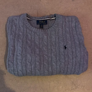Grå kabelstickad tröja Polo Ralph Lauren - Kabelstickad grå tröja från Polo Ralph Lauren med klassisk rund halsringning och broderad logga på bröstet. Tröjan är stickad i mjukt material och har en tidlös look som passar perfekt till en vanlig outfit eller om du vill känna dig lite extra stilig, är det något du undrar så bara skriv/mvh elliot