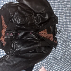 Svart tung skinnjacka i skinnimitation - Säljer en mörkbrun  tjockare jacka i skinnimitation med quiltat brunt innerfoder. Jackan har ribbade muddar, tryckknappar framtill och flera fickor. Perfekt för dig som vill ha en klassisk och stilren look med lite edge. En L men kan funka som XL. Har ett mindre litet hål där back, som knappt syns. 