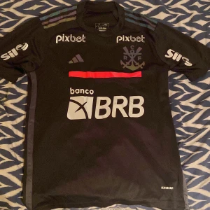 Svart Flamengo fotbollströja Adidas storlek S men passar M - Säljer en svart Flamengo fotbollströja från Adidas med korta ärmar. Tröjan har tryckta sponsorer, klubbmärke på bröstet och klassiska Adidas-ränder på axlarna. Materialet är lätt och andas, perfekt för träning eller match. Slim fit-modell. Den har några skavanker som visas på de två sista bilderna. Annars i bra skick