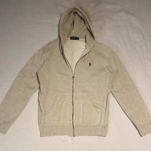 Beige hoodie från Polo Ralph Lauren - Beige hoodie från Polo Ralph Lauren med dragkedja, huva med snörning och klassisk broderad logga på bröstet. Tröjan är sparsamt använd och säljes då jag har växt ur den