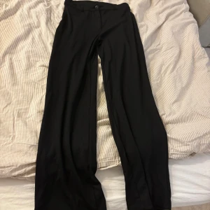 Svarta kostymbyxor från Nelly - Svarta kostymbyxor från Nelly i klassisk rak modell. Byxorna har hög midja, bälteshällor och stängs med knapp och dragkedja. Perfekta för en clean och stilren look. Materialet känns mjukt och följsamt.