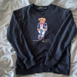 Mörkblå sweatshirt Polo Bear Ralph Lauren - Mörkblå sweatshirt från Polo Ralph Lauren med ikoniska Polo Bear-trycket framtill. Tröjan har rund hals, ribbade muddar och är tillverkad i mjuk bomull. Perfekt för dig som gillar klassisk streetstyle med en twist.