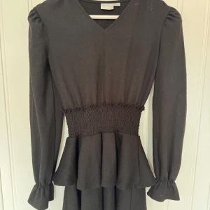 Svart kort klänning från Gina Tricot - Fin svart kort klänning från Gina Tricot med v-ringning, långa puffärmar och smockad midja. Klänningen har volangdetaljer vid midjan. 