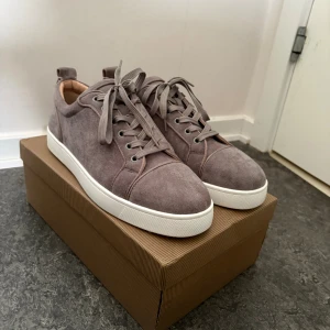 Grå mockasneakers från Louboutin - Snygga sneakers i grå mocka från Christian Louboutin med klassisk röd sula och vita kanter. Skorna har aldrig använts snabb affär= bättre pris