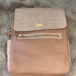 Rosa ryggsäck från DON DONNA - Snygg rosa ryggsäck från MARC JACOBS med guldiga dragkedjor och detaljer. Väskan har en strukturerad yta i skinnimitation och locket har ett ormskinnsmönster. Flera fack och justerbara axelremmar gör den både praktisk och trendig.