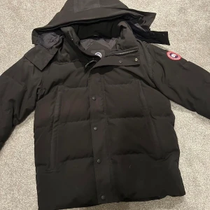 Svart dunjacka från Canada Goose - Säljer en svart dunjacka från Canada Goose med huva och klassisk logga på ärmen. Jackan har dragkedja och knappar framtill, stora fickor och en stilren, puffig design. Perfekt för kalla dagar och riktigt snygg till streetwear-stilen.