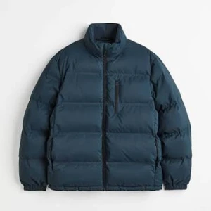 Mörkblå pufferjacka från H&M Man - Snygg mörkblå pufferjacka från H&M Man med hög krage, dragkedja framtill och bröstficka med dragkedja. Jackan har quiltad design, elastiska muddar och tryckknappar vid ärmslut. Perfekt för kalla dagar och enkel att matcha med olika outfits.