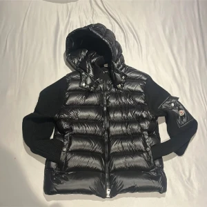 Svart Moncler cardigan  - Passar som L, Snygg svart cardigan från Moncler med glansig quiltad framsida, stickade svarta ärmar och huva. Västen har dragkedja framtill, fickor med tryckknappar och Moncler-logga på ena ärmen. Perfekt för lager-på-lager och streetstyle.