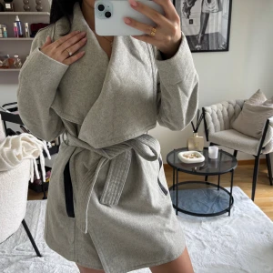 Ljusgrå omlottkappa från Mango XS - Snygg ljusgrå kappa från Mango med omlottmodell och brett slag. Jackan har knytskärp i midjan och svarta detaljer vid fickorna. Perfekt för dig som gillar stilrena och trendiga plagg med en relaxed vibe.