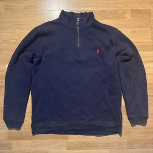 Mörkblå half zip tröja Polo Ralph Lauren - Mörkblå långärmad tröja från Polo Ralph Lauren med klassisk röd logga broderad på bröstet. Tröjan har en hög krage med half zip-dragkedja och är tillverkad i mjuk bomull. Perfekt för lager-på-lager och chill stil.