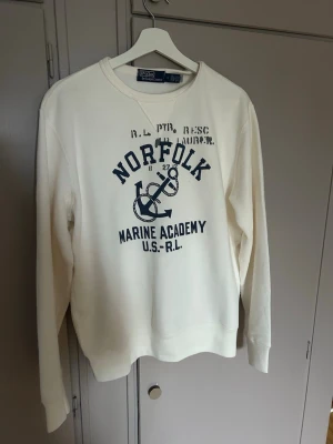 Vit sweatshirt Polo Ralph Lauren M - Säljer en vit sweatshirt från Polo Ralph Lauren i storlek M. Tröjan har marinblå tryck med texten 'Norfolk Marine Academy' och ankare på framsidan samt tryck på ryggen. Klassisk rund hals och långa ärmar. Perfekt för en avslappnad stil. Använda 1 gång 10/10 skick