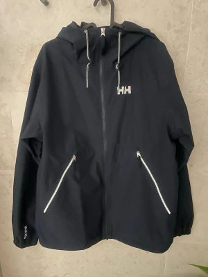 Svart vindjacka från Helly Hansen - Säljer en svart vindjacka från Helly Hansen med huva och vita dragkedjor. Jackan har två snedställda fickor framtill, justerbar huva med snören och logga på bröstet samt ryggen. Perfekt för blåsiga dagar och enkel att matcha med allt.