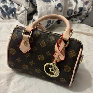 Brun handväska med mönster och gulddetaljer - Snygg brun handväska med guldmönster och ljusa handtag. Väskan har guldfärgade metalldetaljer, dragkedja och ett dekorativt hänge framtill. Insidan är randig och det finns en avtagbar axelrem. Perfekt för dig som gillar stilrena accessoarer med lyxig känsla.