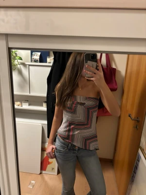 Randig bandeau tubtopp från Muaa - Säljer en bandeau tubtopp från Muaa med grafiskt randigt mönster i svart, vitt och rött. Toppen är axelbandslös och har en liten ringdetalj framtill. Materialet känns lätt och svalt, perfekt för varma dagar.