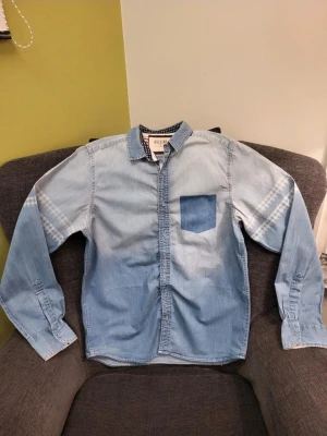 Ljusblå jeansskjorta från Big Star M - Säljer en ljusblå jeansskjorta från Big Star med lång ärm och en bröstficka. Insidan av kragen har stjärnmönster och manschetterna har röda och vita ränder. Skjortan har knappar framtill och coola rutiga detaljer på ärmarna.