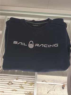 Mörkblå Sail Racing sweatshirt - Mörkblå sweatshirt från Sail Racing med vit logga tryckt över bröstet. Klassisk rund halsringning och ribbade muddar vid ärmslut och nederkant. Tillverkad i mjuk bomull, perfekt för chill dagar eller när du vill ha en clean look.